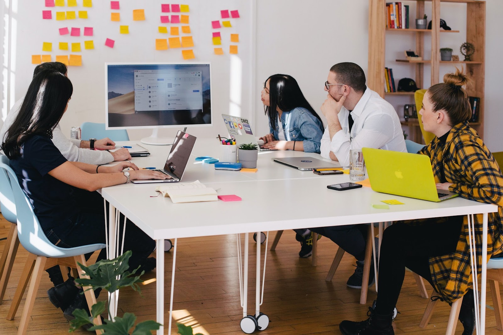 A team of five employees sit at a white table and look at a computer screen at the end. A whiteboard in the background is covered with yellow and pink notes for tasks.