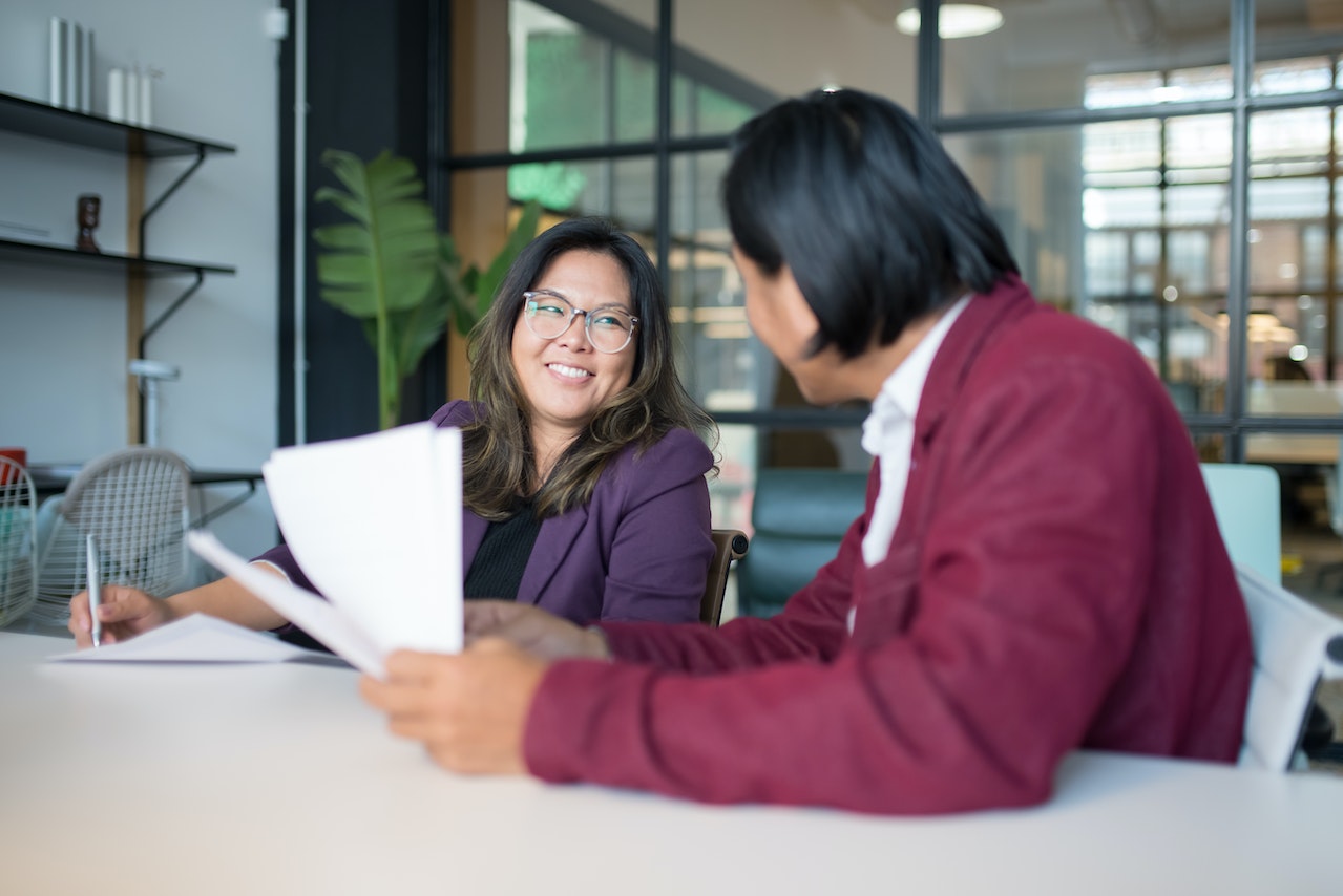 Photo of two employees, one smiling at the other, sitting at a table and holding papers