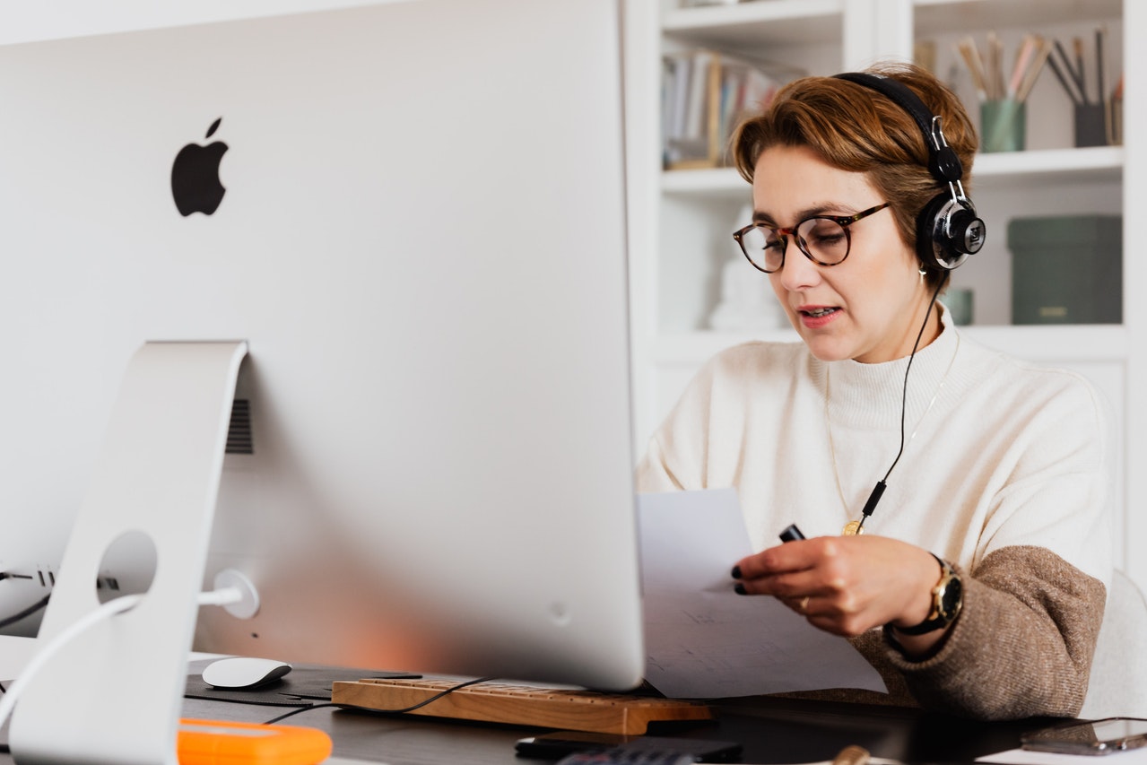 A woman is sitting in front of a computer and wearing a heatset. She looks at a piece of paper and holds a pen in her hand