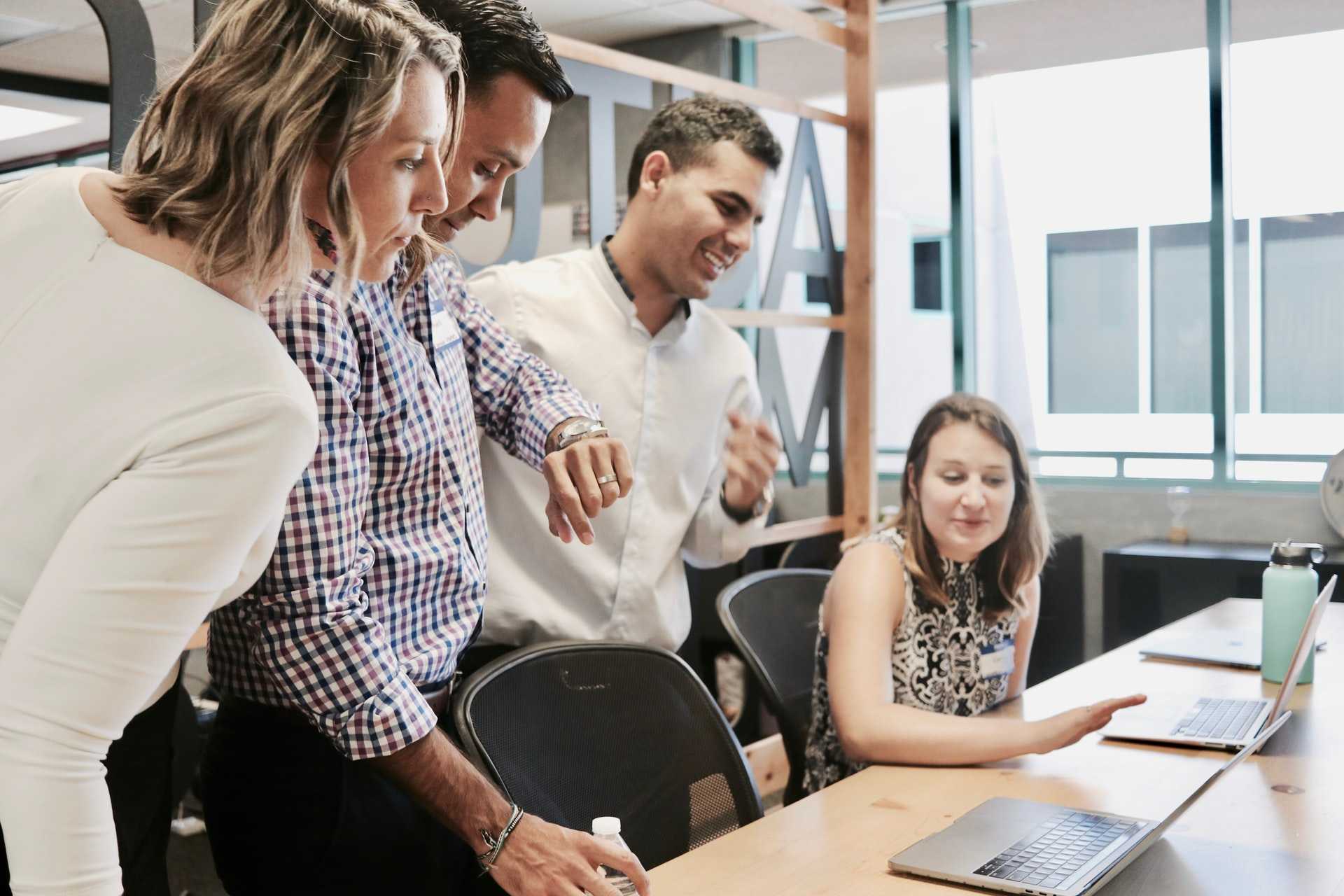 Photo of four employees, two men and a woman standing up, and a woman sitting down at a meeting table looking at a laptop on the table