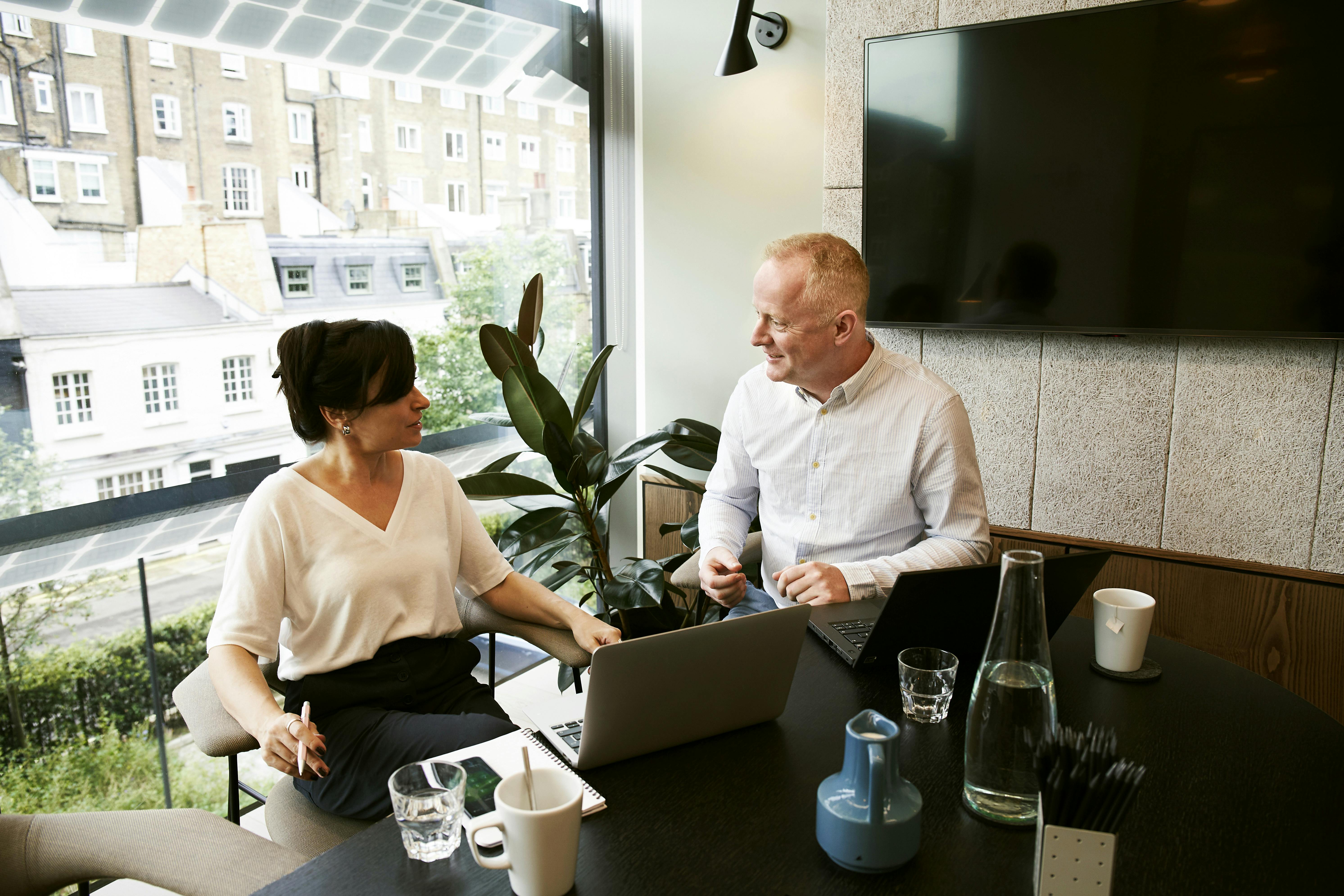 A photo of two professionals working together in an informal meeting space next to a large window.