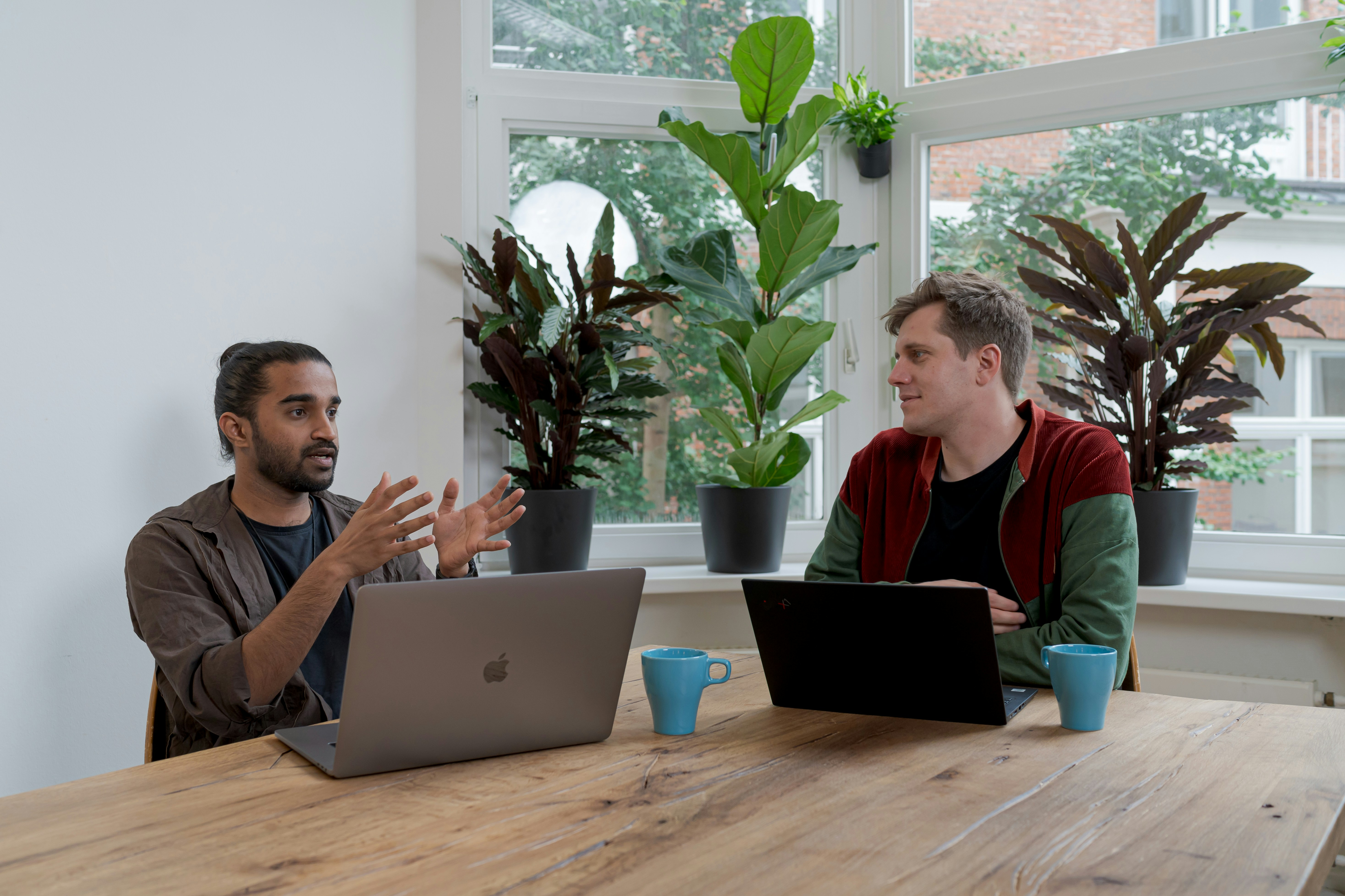 A photo of two professionals sitting at a large table together with their laptops out, having a discussion.